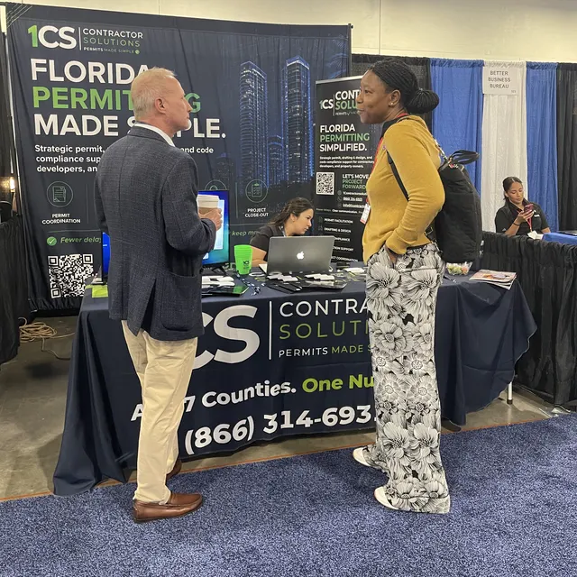 Two attendees talking in front of the 1CS booth, with the Florida Permitting Made Simple banner behind them